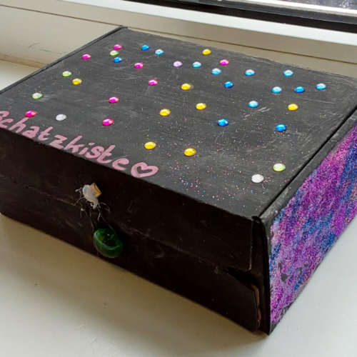 Die geschlossene Wertschätzungskiste ist schwarz, lila und blau und mit Glitzersteinchen beklebt. Auf dem Deckel steht in rosa Schrift "Schatzkiste" mit einem Herz dahinter.