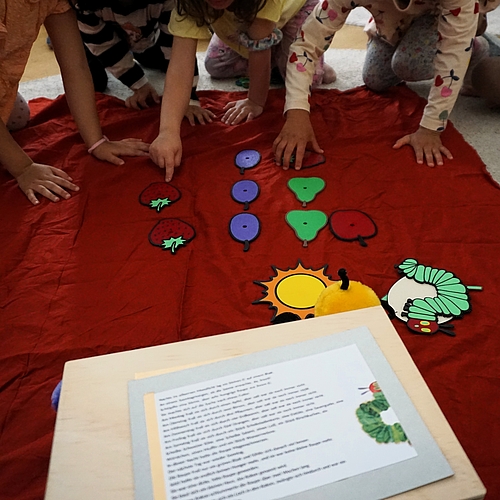 Kinder sitzen um eine rote Stoffunterlage und ordnen bunte Bildkarten mit Obst, Sonne und Raupe